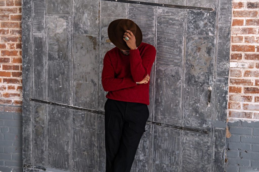 Stylish man leaning against a textured wall, wearing a brown hat and red sweater.
